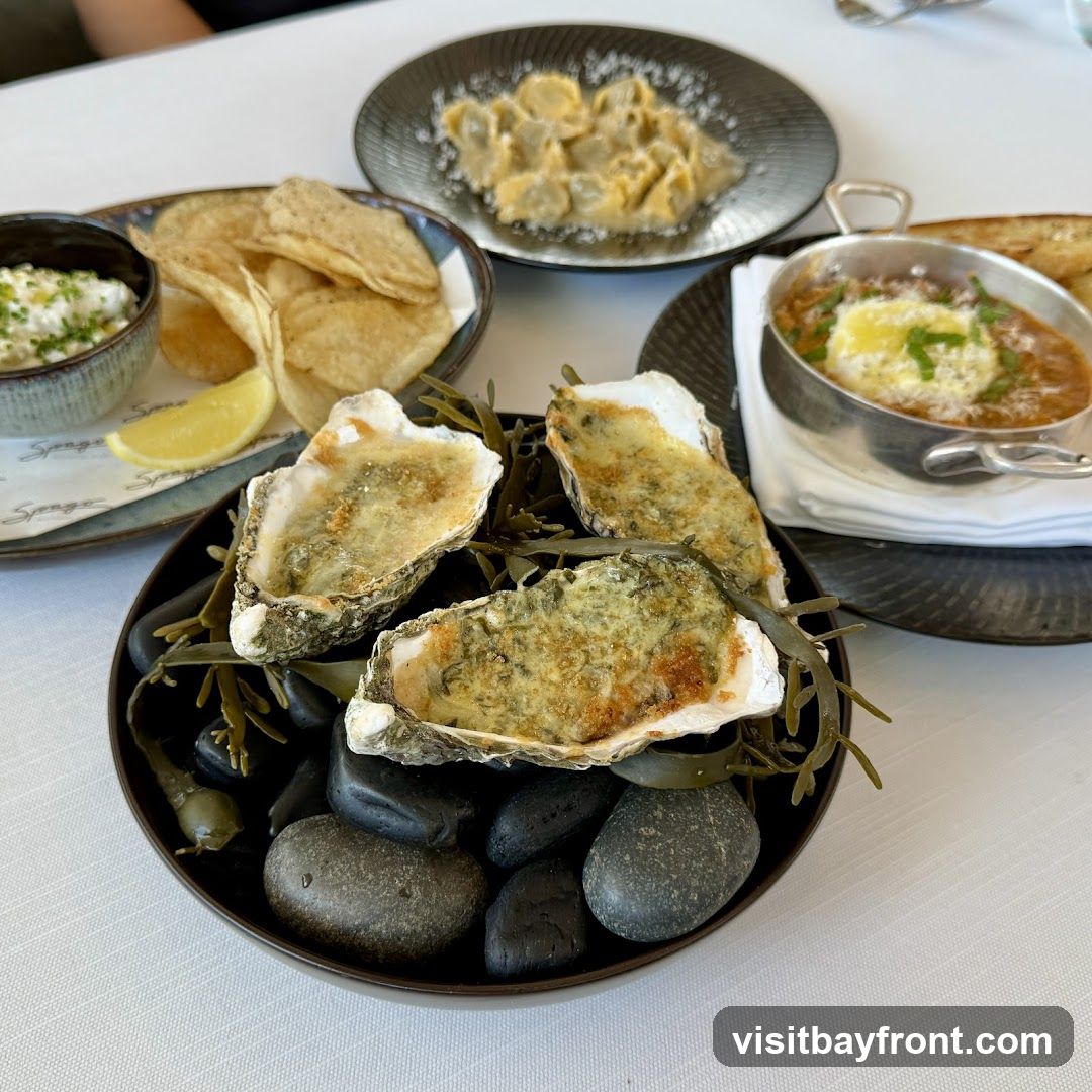 A close-up shot of a table setting with several dishes. In the foreground, a dark bowl holds three baked oysters on a bed of seaweed and dark stones. To the left, a small bowl contains a creamy dip with chives, accompanied by potato chips and a lemon wedge. Behind the oysters, a plate of tortellini is visible, and to the right, a small metal dish holds a stew-like dish topped with cheese and herbs, with toast on the side. The table is covered with a white tablecloth. A logo that appears to say "Spago" is visible on a paper liner under the chips and dip.