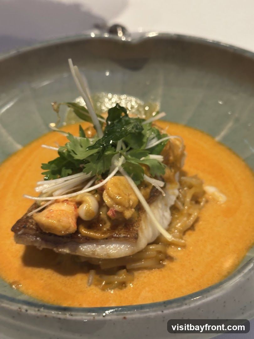 A close-up shot of a piece of white fish served in a shallow bowl with a vibrant orange curry sauce. The fish is topped with what appears to be small pieces of lobster or shrimp, and garnished with fresh cilantro and thinly sliced white vegetables. The dish is presented on a light-colored surface.