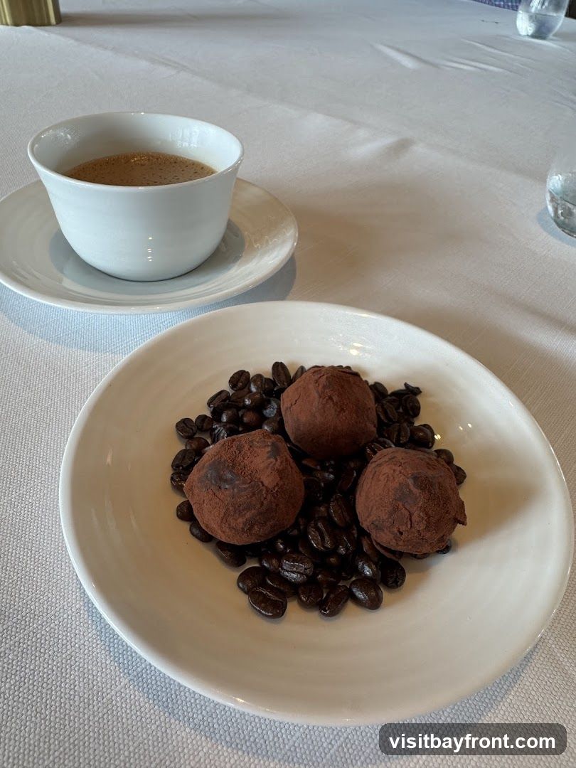 A white cup of coffee sits on a saucer next to a white plate filled with coffee beans and three chocolate truffles dusted with cocoa powder. The setting appears to be a table with a white tablecloth.