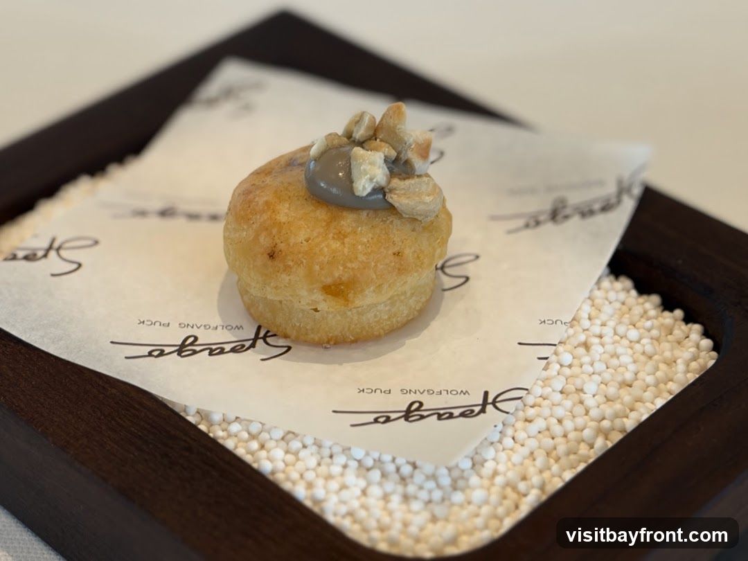 A small, golden-brown pastry sits on a piece of white paper with a black logo. The pastry is topped with a dollop of grey cream and chopped nuts. The paper is placed in a dark wooden tray filled with small white beads.
