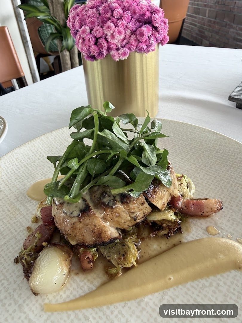A close-up shot of a plated dish featuring grilled chicken, roasted vegetables, and a creamy sauce. The dish is garnished with fresh greens and served on a textured white plate. A golden vase with pink flowers sits in the background, along with a glimpse of a restaurant interior.