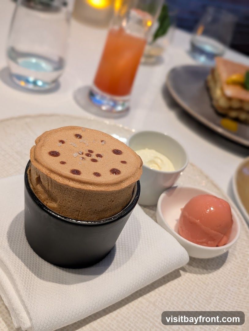 A close-up of a chocolate soufflé served in a black ramekin, topped with sea salt and small dots of what appears to be chocolate or fruit. Beside the soufflé are two small white bowls, one containing a scoop of pink sorbet and the other a dollop of cream. In the background, out of focus, are a glass of water, a tall glass with a pink beverage, and a dessert on a plate.