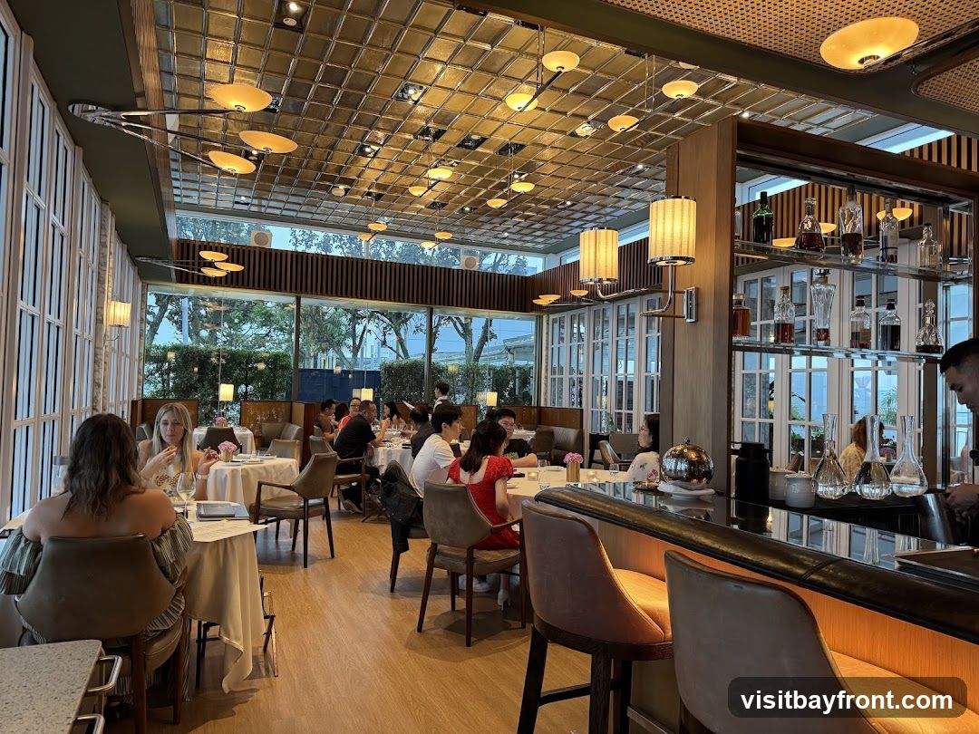 The image shows the interior of a restaurant with a bar area in the foreground and dining tables in the background. The bar has illuminated seating and a reflective countertop. Behind the bar, shelves are stocked with various bottles and glassware. The dining area features tables set with white tablecloths, and patrons are seated and dining. Large windows line the back wall, offering a view of greenery outside. The ceiling is adorned with a grid of square panels and decorative lighting fixtures.