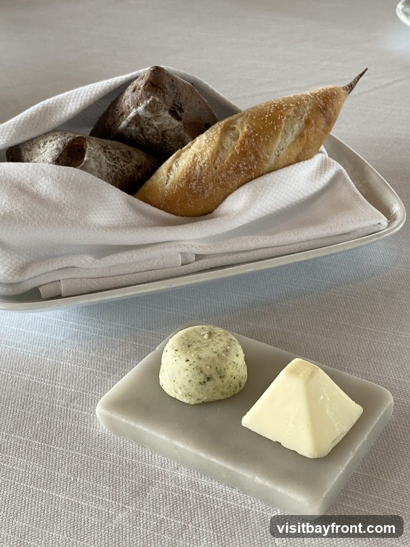 A white cloth-lined basket holds an assortment of bread, including a baguette and two round loaves. In the foreground, a small grey dish holds two types of butter: one is a round pat of herb butter, and the other is a triangular piece of plain butter. The setting appears to be a restaurant table with a white tablecloth.