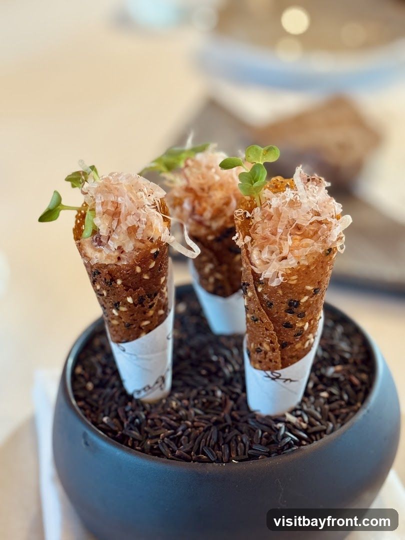 Three small, crispy cones filled with a savory mixture and topped with bonito flakes and microgreens. The cones are dark brown and appear to be made with sesame seeds. They are presented in a black bowl filled with black rice.