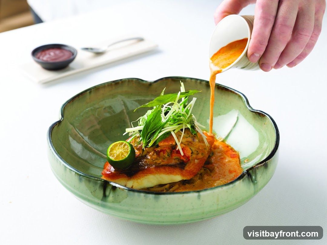 A hand is pouring a rich orange sauce over a piece of fish in a green, lotus-shaped bowl. The fish is topped with fresh green herbs and a slice of lime. A small dish of dark red sauce and a spoon are visible in the background.