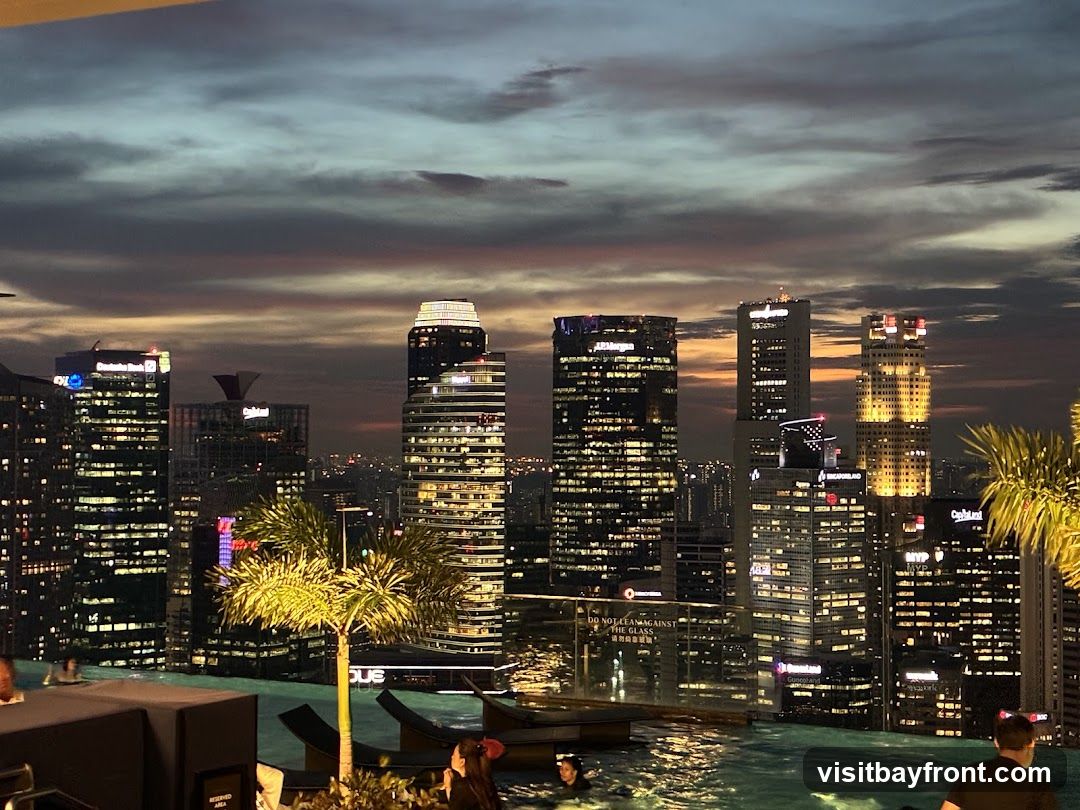 An infinity pool with people relaxing in it is in the foreground, with lounge chairs visible. Beyond the pool, a sprawling cityscape of illuminated skyscrapers stretches into the distance under a dramatic twilight sky with hues of orange, pink, and gray. The lights of the city buildings create a vibrant contrast against the darkening sky.
