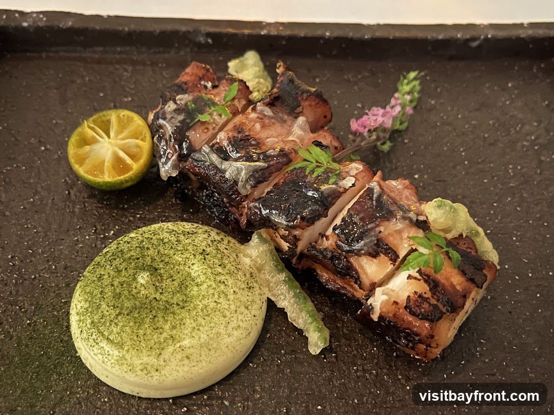 A close-up shot of a grilled dish served on a dark textured plate. The dish appears to be sliced meat with char marks, garnished with fresh green herbs and a small sprig of pink flowers. To the left, a slice of lime and a dollop of creamy green-dusted sauce are visible.