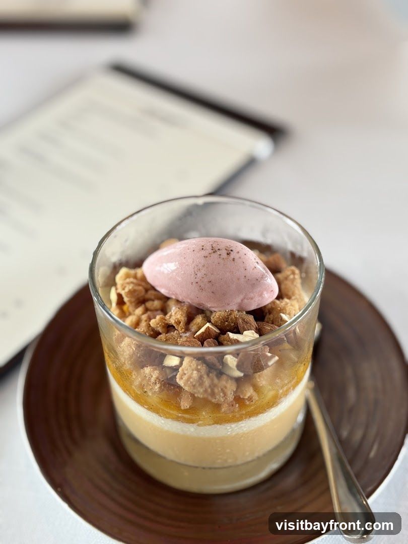 A layered dessert in a glass, topped with a scoop of pink sorbet and crumbled nuts. A menu is visible in the blurred background.