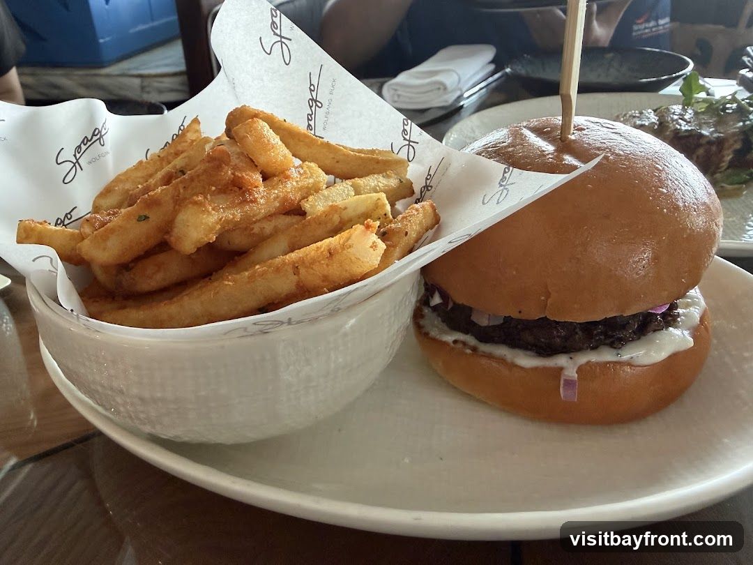 A close-up shot of a burger and a bowl of fries. The burger is on a white plate with a wooden skewer sticking out of the top bun. The fries are in a white bowl lined with paper that has the Spago Wolfgang Puck logo.