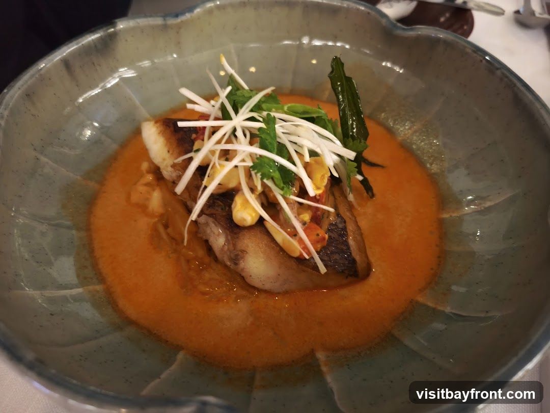 A close-up shot of a piece of fish served in a shallow bowl with a vibrant orange sauce. The fish is topped with thinly sliced white scallions, cilantro, and what appears to be corn kernels and diced red peppers. A single green chili pepper is also visible. The bowl has a textured, grey-blue glaze with a scalloped edge.
