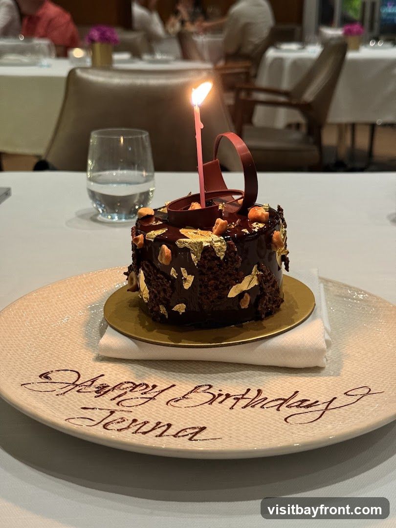 A small, dark chocolate cake with a lit candle sits on a plate. The cake is decorated with chocolate icing, nuts, and gold leaf. The plate has "Happy Birthday Jenna" written on it in chocolate sauce. The setting appears to be a restaurant with tables and chairs in the background.