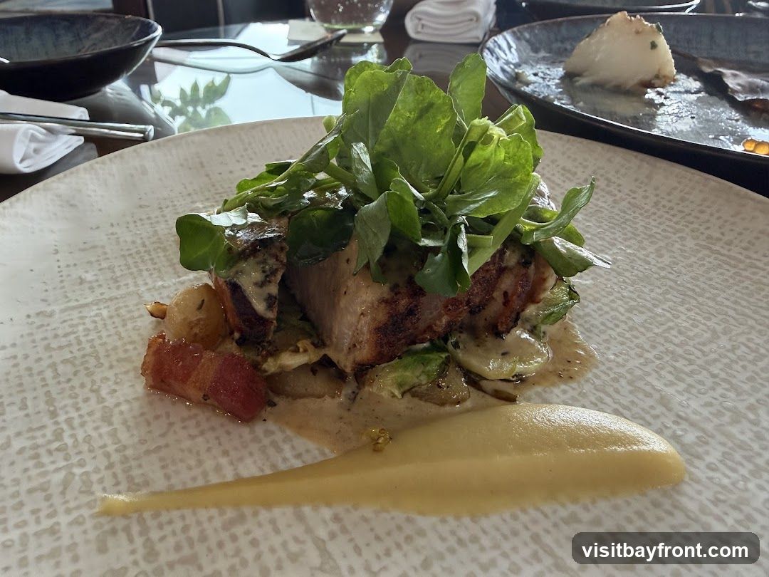 A close-up shot of a gourmet dish featuring pork belly, garnished with fresh greens and served with a creamy sauce and a dollop of puree. The plate has a textured white surface. In the background, partially visible, are other dining elements like bowls, cutlery, and a partially eaten dish on another plate.