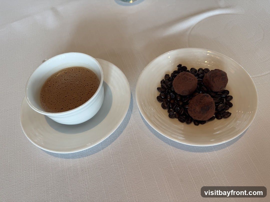 A white cup of coffee with foam sits on a saucer next to a white bowl filled with coffee beans and three chocolate truffles.