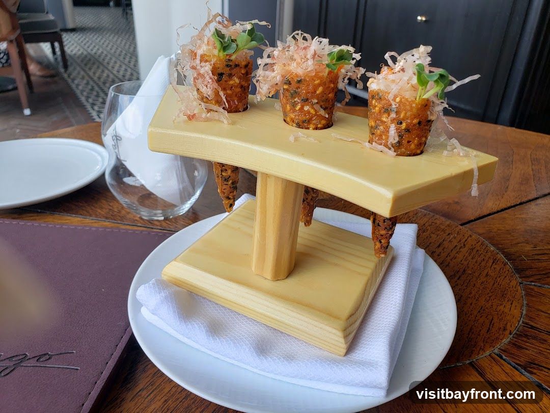 Three small appetizers, possibly sushi rolls or rice cones, are presented on a light-colored wooden stand. Each appetizer is topped with microgreens and garnished with what appears to be dried bonito flakes. The appetizers are served on a white plate with a white napkin. A wine glass and another plate are visible in the background, along with a dark purple menu cover. The setting appears to be a restaurant with a wooden table.