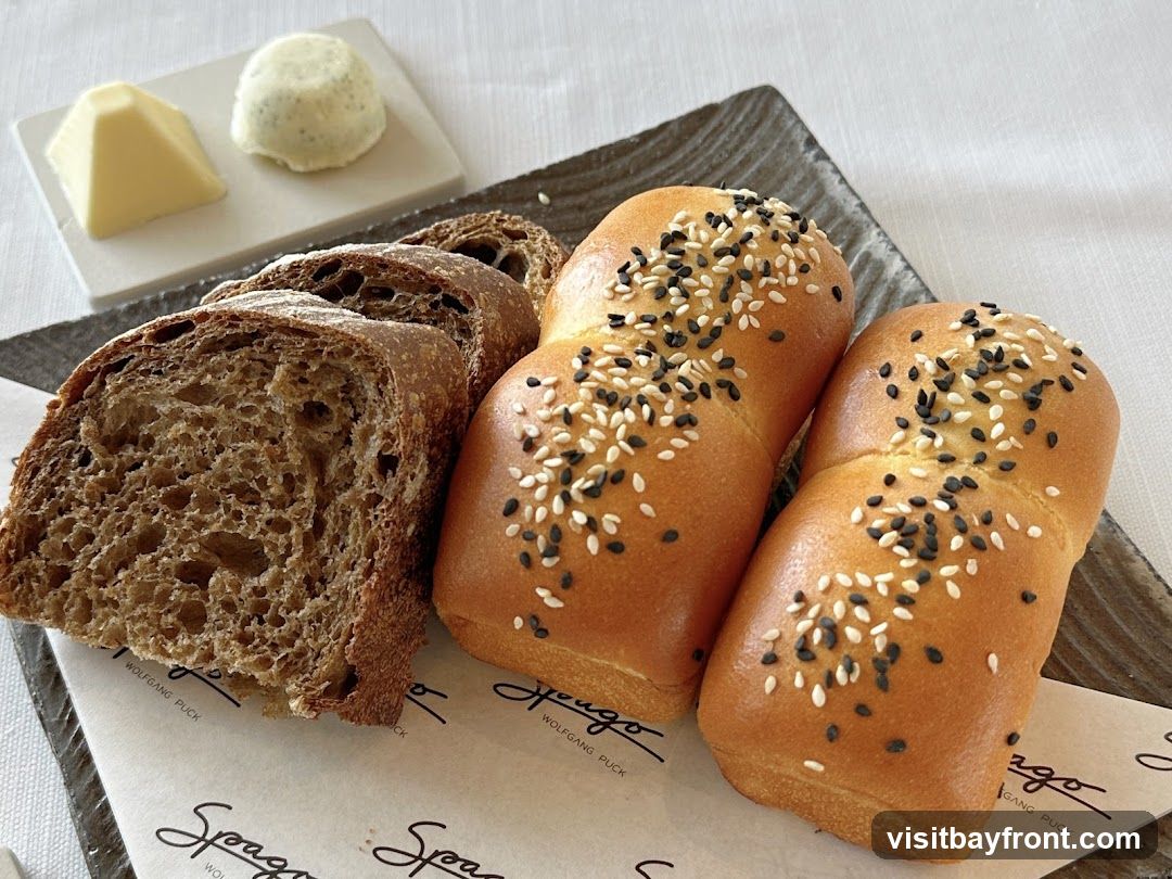 A close-up shot of a bread basket containing sliced dark bread and two rolls topped with sesame seeds. A small dish with two portions of butter is visible in the background. The bread is served on a dark rectangular plate with a paper liner that has the Wolfgang Puck logo printed on it.