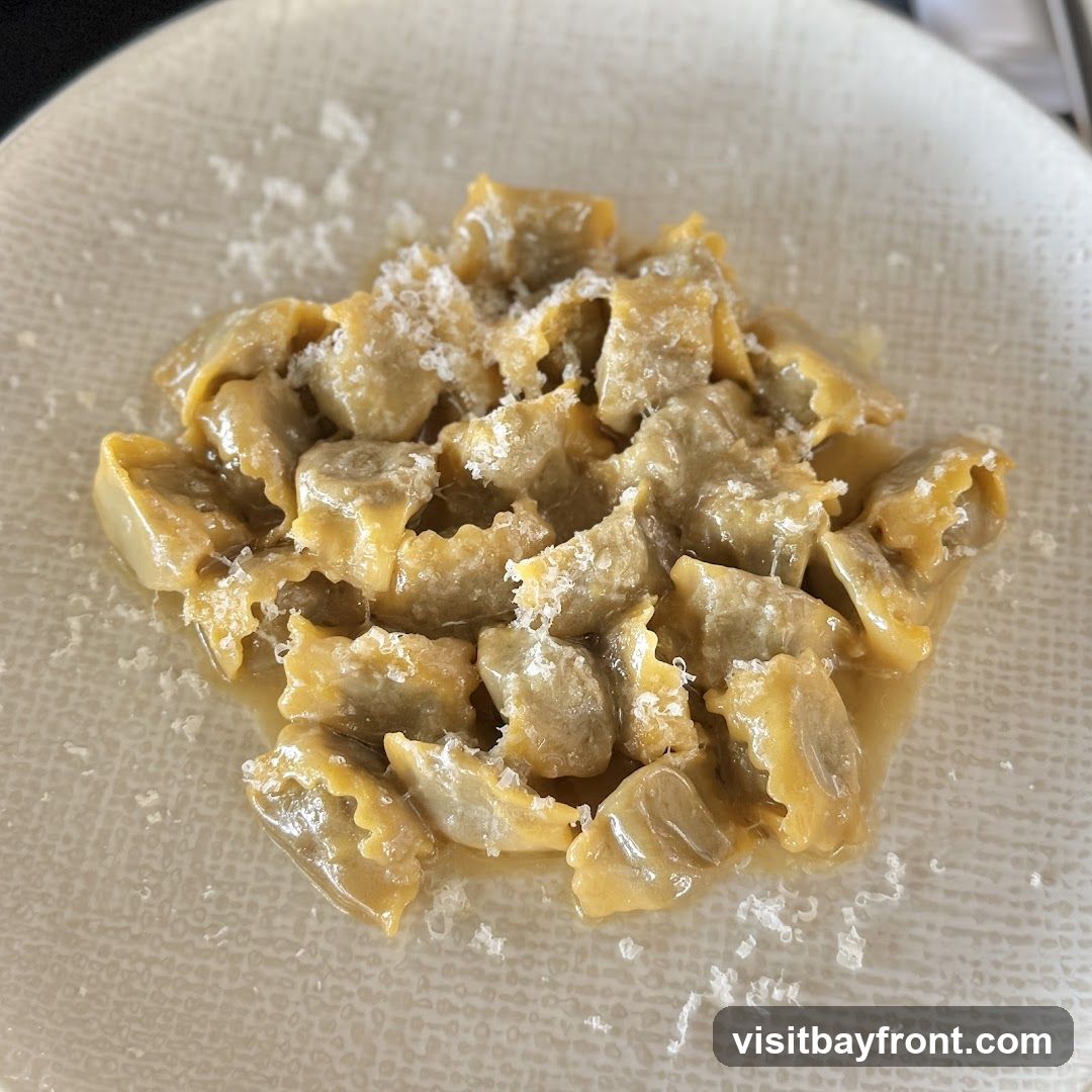 A close-up shot of a plate of pasta, likely tortellini or ravioli, served in a light sauce and topped with grated cheese. The pasta pieces are small and folded, with visible crimped edges. The dish is presented on a textured, light-colored plate.
