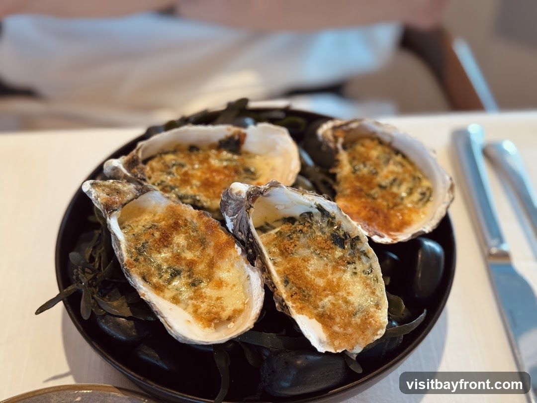Four baked oysters are served on a black plate with dark seaweed and smooth stones. The oysters are topped with a golden-brown breadcrumb and cheese mixture, with visible green herbs mixed in. The dish is presented on a white tablecloth, with cutlery visible to the right. The background is blurred, showing a person in a white shirt and a hint of a wooden chair.