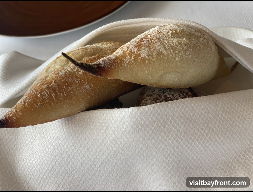 A close-up shot of a bread basket filled with several loaves of bread, some dusted with flour. The bread is wrapped in a white cloth napkin. A dark-rimmed plate is visible in the upper left corner.