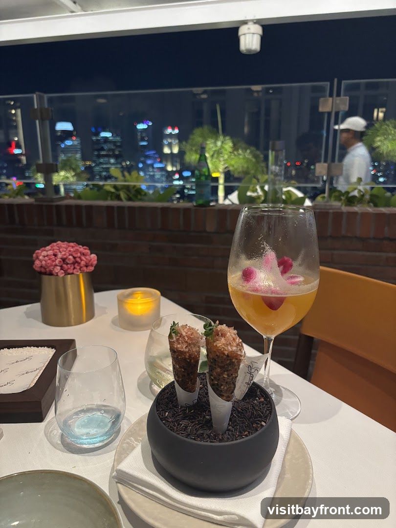 A table set for dining on a rooftop at night, with a cityscape visible in the background. On the table, there are two small appetizers served in cones, a cocktail garnished with flowers, a candle, and glassware. A person in a white hat is visible in the background.