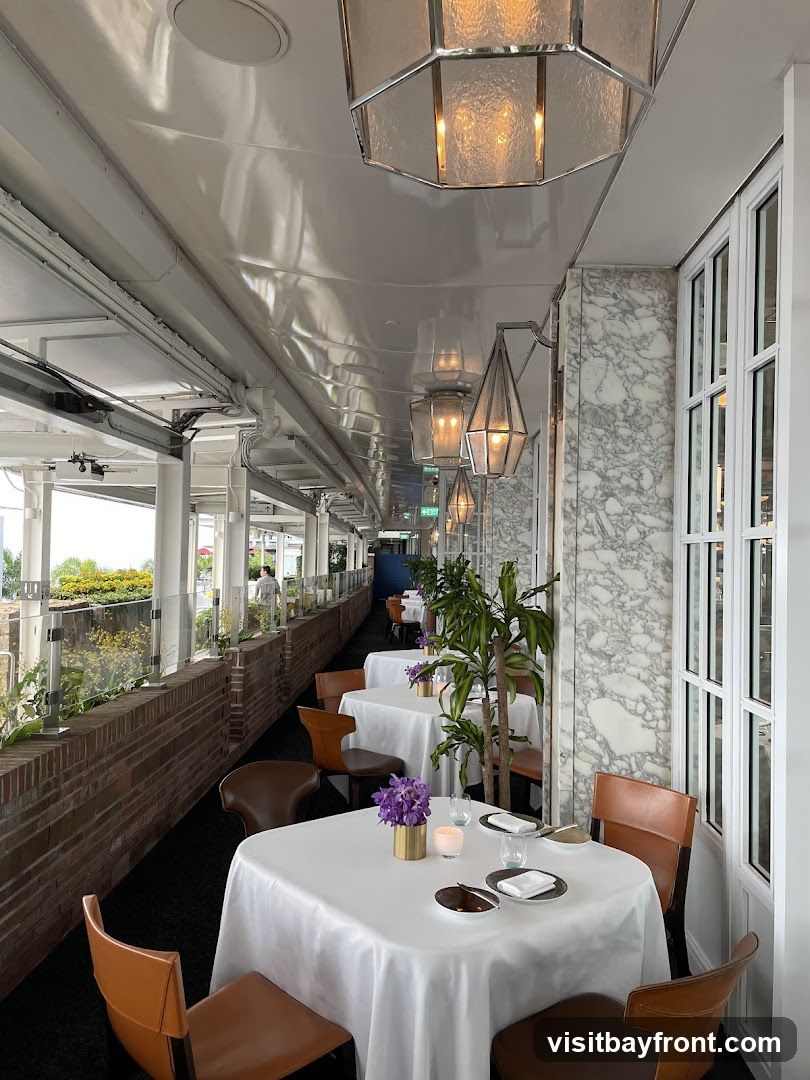 An elegant outdoor dining area with tables set for a meal. White tablecloths cover square tables, each adorned with a small vase of purple flowers, a candle, and place settings. Brown leather chairs surround the tables. The area features marble-patterned walls, large windows, and decorative pendant lights hanging from the ceiling. A brick planter with greenery runs along the side, and in the distance, more tables and a few patrons are visible.