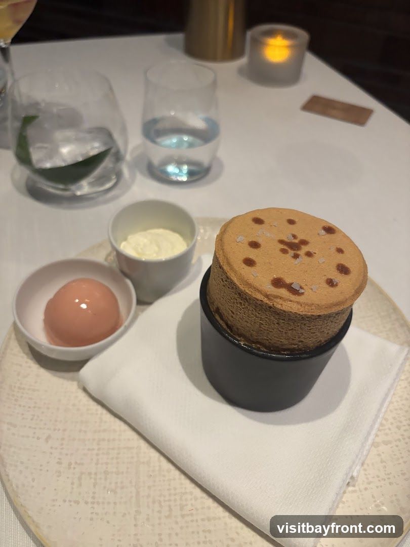 A close-up shot of a dessert featuring a soufflé, a scoop of pink sorbet, and a small dish of cream, all presented on a textured plate with a white napkin. Two glasses of water and a lit candle are visible in the background, suggesting a dining setting.