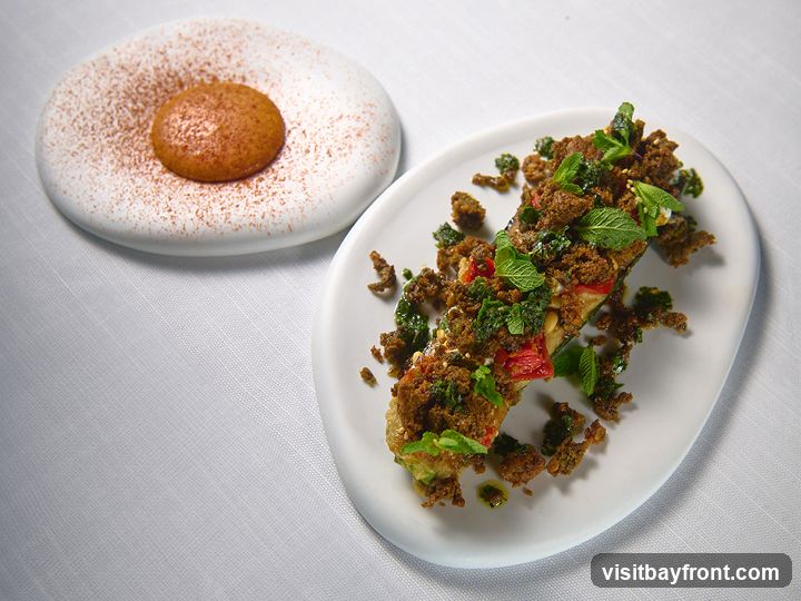 A close-up shot of two artfully presented dishes on a white tablecloth. The dish in the foreground is a long, halved vegetable, possibly eggplant, topped with a crumbly mixture, fresh mint leaves, and a drizzle of sauce. The dish in the background is a round, shallow plate with a single, golden-brown sphere dusted with a reddish-brown powder.