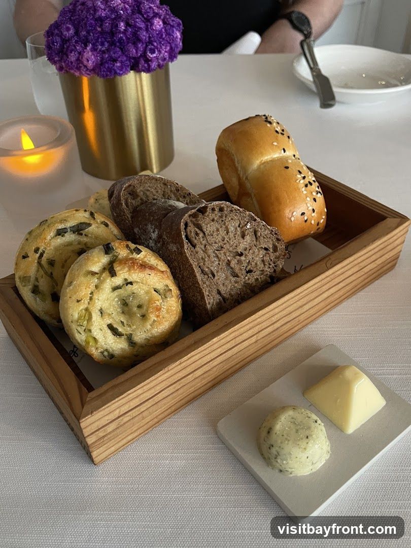 A wooden basket filled with assorted bread rolls, including a plain roll topped with sesame seeds, a dark rye bread, and a green onion swirl bread. A small dish holds two types of butter: a round dollop of herb butter and a triangular pat of plain butter. A lit candle and a vase with purple flowers are visible in the background.
