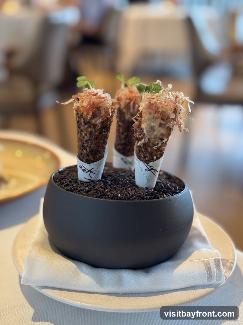 Three appetizer cones filled with a savory mixture and topped with shredded fish and microgreens, presented in a black bowl filled with black rice. The cones have paper wrappers with a logo and text. The background shows a blurred restaurant interior with tables and chairs.