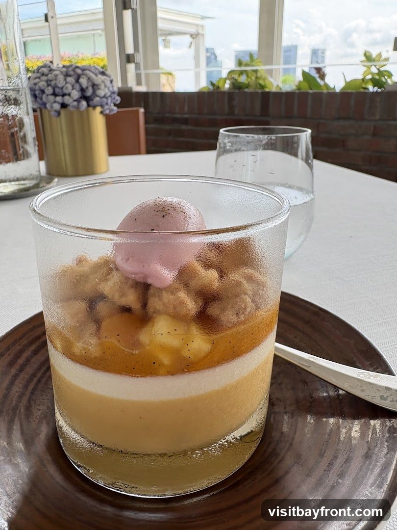 A layered dessert in a glass, topped with a scoop of pink ice cream and crumble. The dessert appears to have layers of custard or mousse. It is served on a dark plate with a fork. In the background, there is a vase with purple flowers, a glass of water, and a window with a view of buildings and greenery.