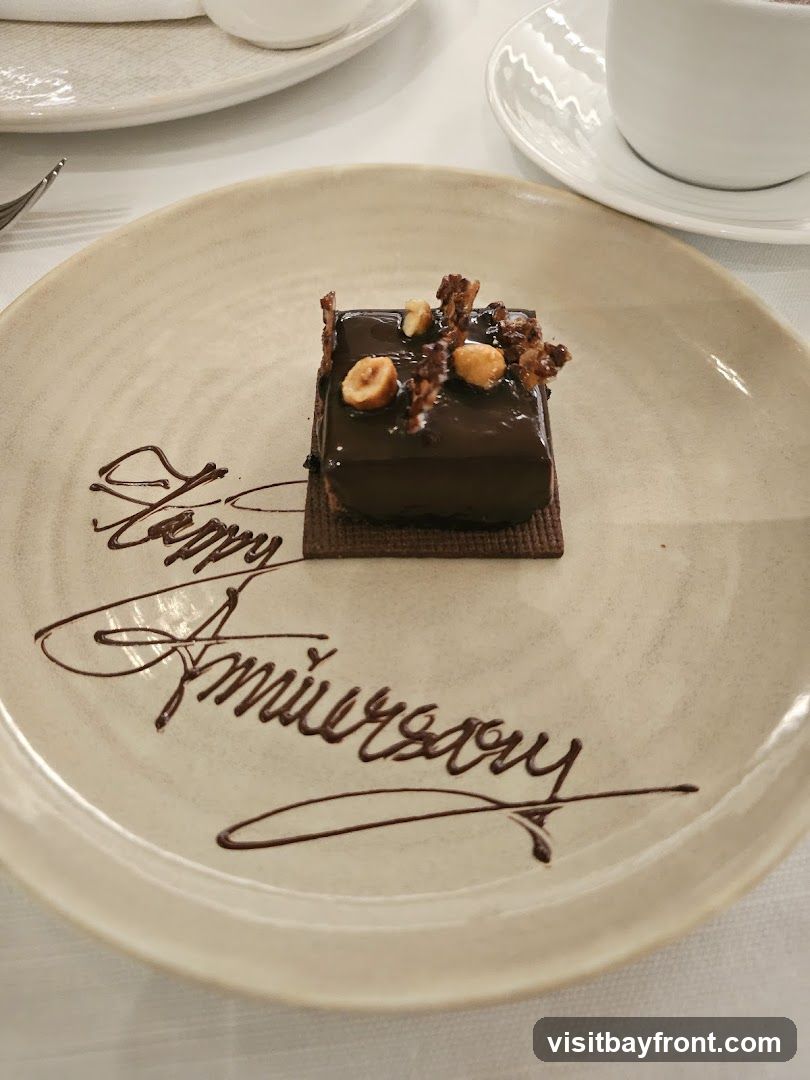 A small, square chocolate dessert topped with nuts and caramel shards sits on a plate. The plate is decorated with chocolate writing that says "Happy Anniversary". A cup and saucer are visible in the background.