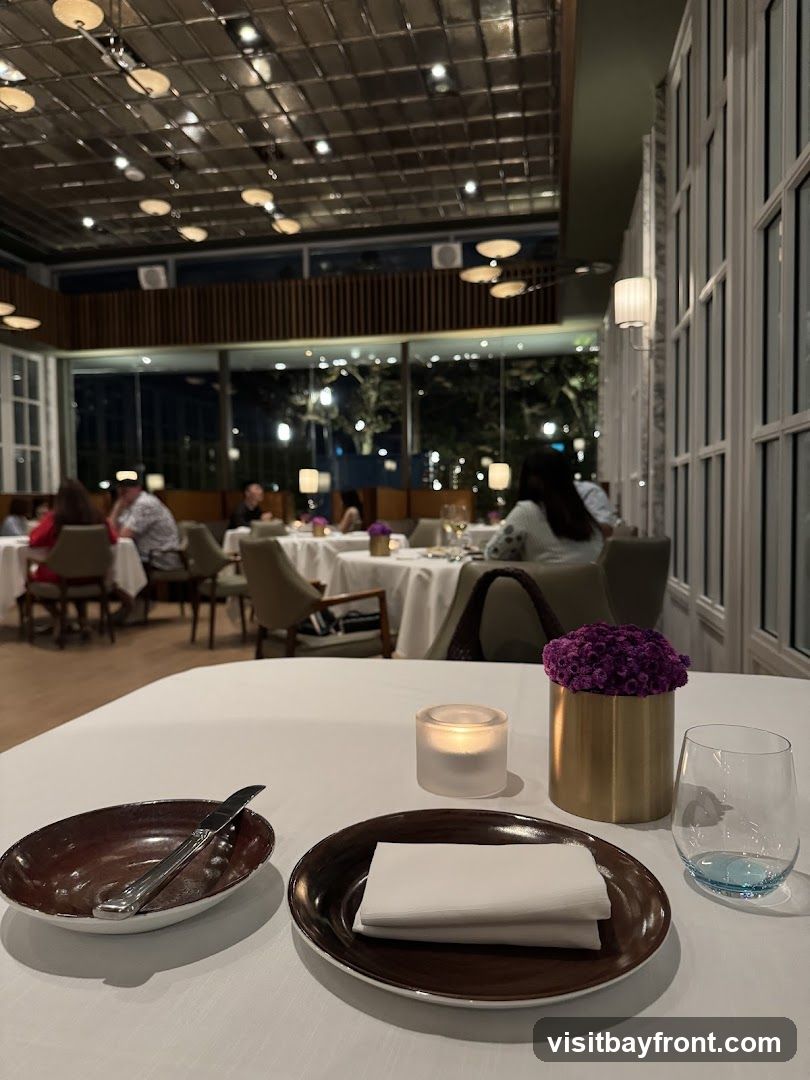 A close-up view of a dining table set for a meal in an upscale restaurant. The table is covered with a white tablecloth. In the foreground, there is a dark brown oval dish with a knife, a smaller dark brown plate with folded white napkins, a lit candle in a frosted glass holder, a golden vase with a vibrant purple floral arrangement, and a clear wine glass with a light blue base. In the background, other tables are set with white tablecloths, and diners are visible, suggesting a lively yet refined atmosphere. The restaurant features a modern ceiling with numerous lights and large windows overlooking an outdoor area at night.