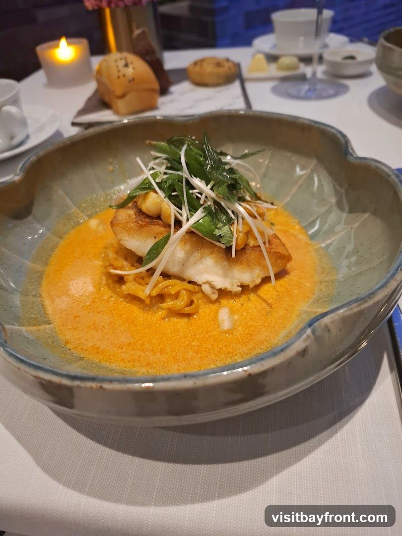 A close-up shot of a fish dish served in a decorative bowl. The fish is topped with green onions and other garnishes, and sits in a vibrant orange sauce with noodles. In the background, a lit candle, bread rolls, and a wine glass are visible on a white tablecloth.