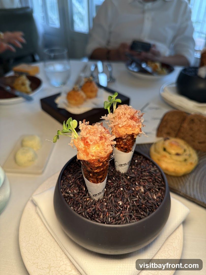 Two small appetizer cones filled with a creamy mixture and topped with shredded ingredients and microgreens are presented in a bowl of black rice. In the background, other small bites, bread, and drinks are visible on a table setting with diners.