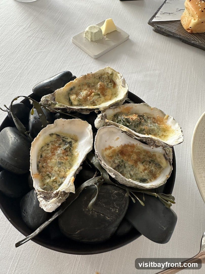 A close-up shot of four baked oysters served in their shells on a bed of dark stones. The oysters are topped with a creamy sauce and breadcrumbs, and appear to be garnished with herbs. In the background, a small plate holds a cube of butter and a wedge of cheese, and a piece of bread is visible on a dark platter. The table is covered with a white tablecloth.