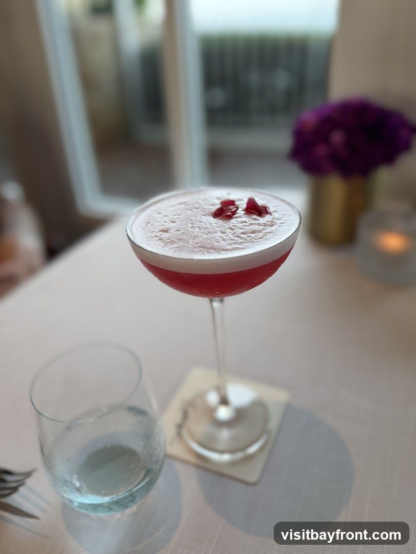 A close-up shot of a pink cocktail in a coupe glass, garnished with two small red flower petals. The drink has a frothy top. Next to it is a glass of water and a coaster. In the background, a vase with purple flowers and a lit candle are visible. The setting appears to be a restaurant or bar with a white tablecloth.