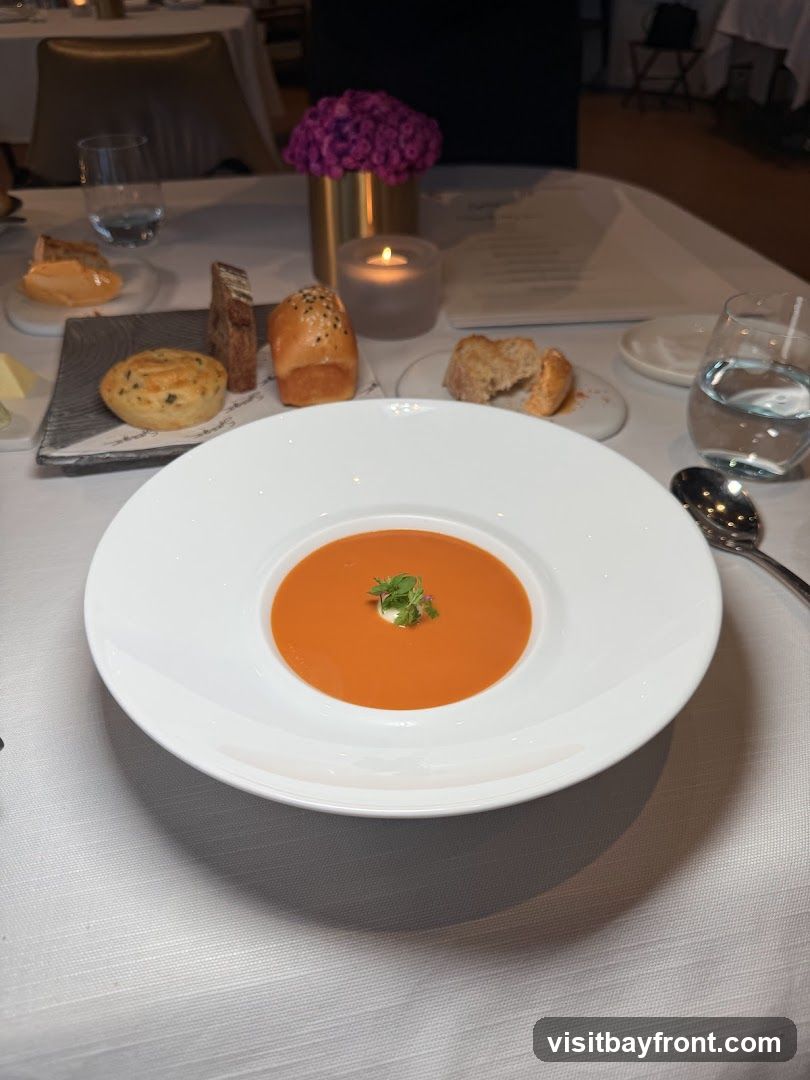 A white bowl of tomato soup with a dollop of cream and a sprig of garnish is centered on a table set with a white tablecloth. To the left, a small plate holds a savory muffin, a slice of bread, and a small bun with sesame seeds. Another small plate to the right has broken pieces of bread. A lit candle in a glass holder and a vase of purple flowers are visible in the background, along with a menu.