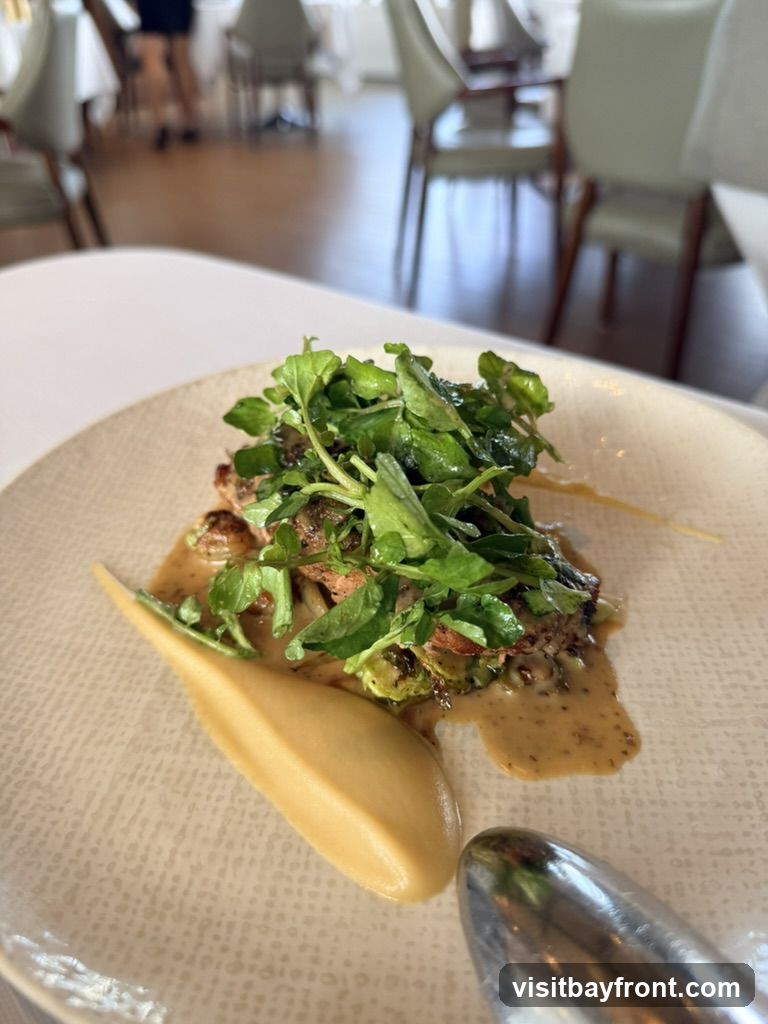 A close-up shot of a finely plated dish featuring a piece of meat topped with fresh green leaves, served with a smooth, golden puree and a rich sauce. The plate has a textured pattern. In the blurred background, a restaurant interior with tables and chairs is visible.