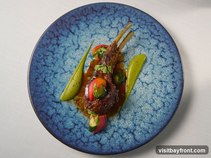 A beautifully plated rack of lamb is presented on a textured blue plate. The lamb chops are coated in a rich, dark sauce and garnished with vibrant green puree, sliced tomatoes, and fresh mint leaves. The dish is artfully arranged with a side of what appears to be zucchini and red pepper.