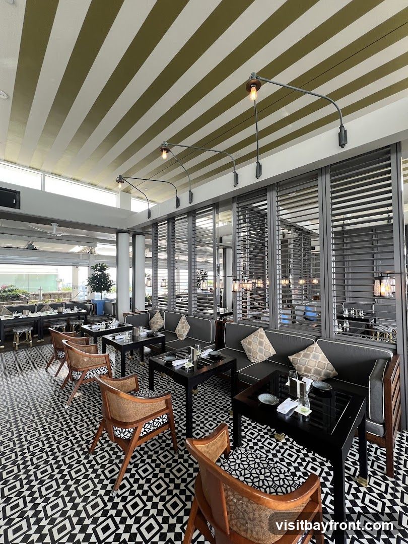 A well-lit restaurant interior with a striking black and white patterned floor. The seating area features dark wood tables and chairs with woven backs, complemented by plush grey banquettes with decorative pillows. The ceiling is adorned with alternating stripes of white and olive green, and industrial-style pendant lights hang from metal arms. Large louvered windows or dividers create a sense of depth and privacy. In the background, more tables are set for dining, and a glimpse of an outdoor area with greenery is visible.