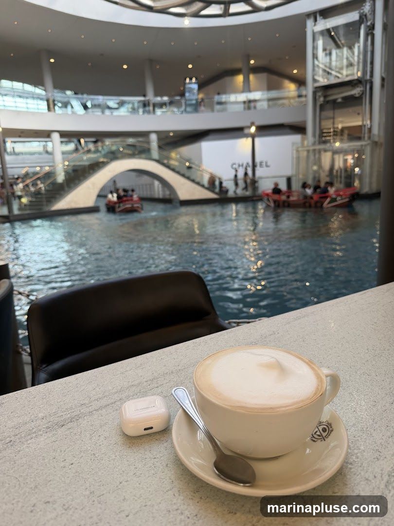 A cup of latte with foam art sits on a saucer with a spoon, next to a pair of white wireless earbuds on a counter. In the background, a canal with boats and people is visible, with a multi-level shopping mall featuring escalators and a Chanel store in the distance.