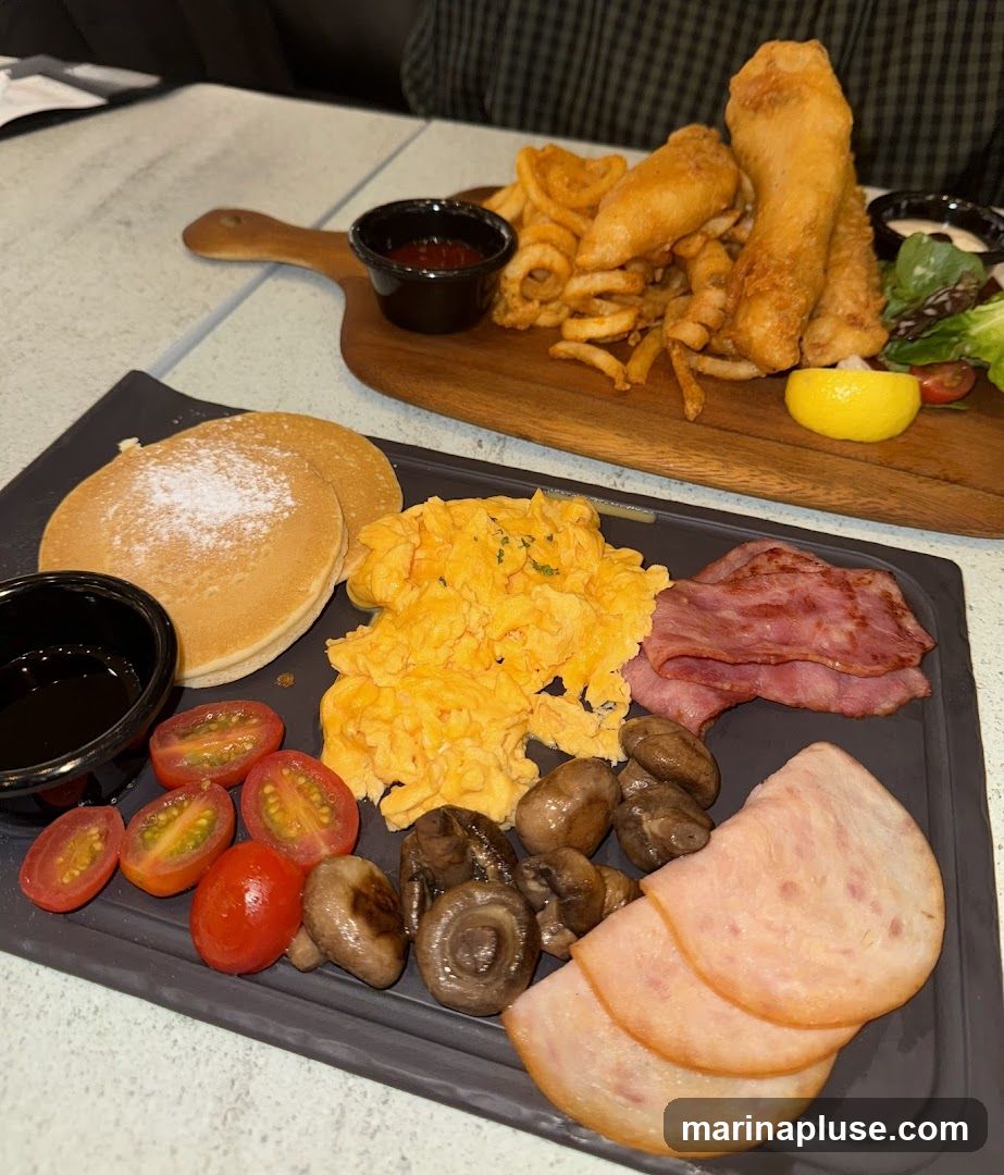 A close-up shot of two breakfast platters. The platter in the foreground features pancakes dusted with powdered sugar, scrambled eggs, sliced cherry tomatoes, sautéed mushrooms, and ham. A small bowl of syrup is to the left. The platter in the background, served on a wooden board, contains fish and chips with curly fries, a small salad, and a lemon wedge. A small bowl of ketchup is also visible.