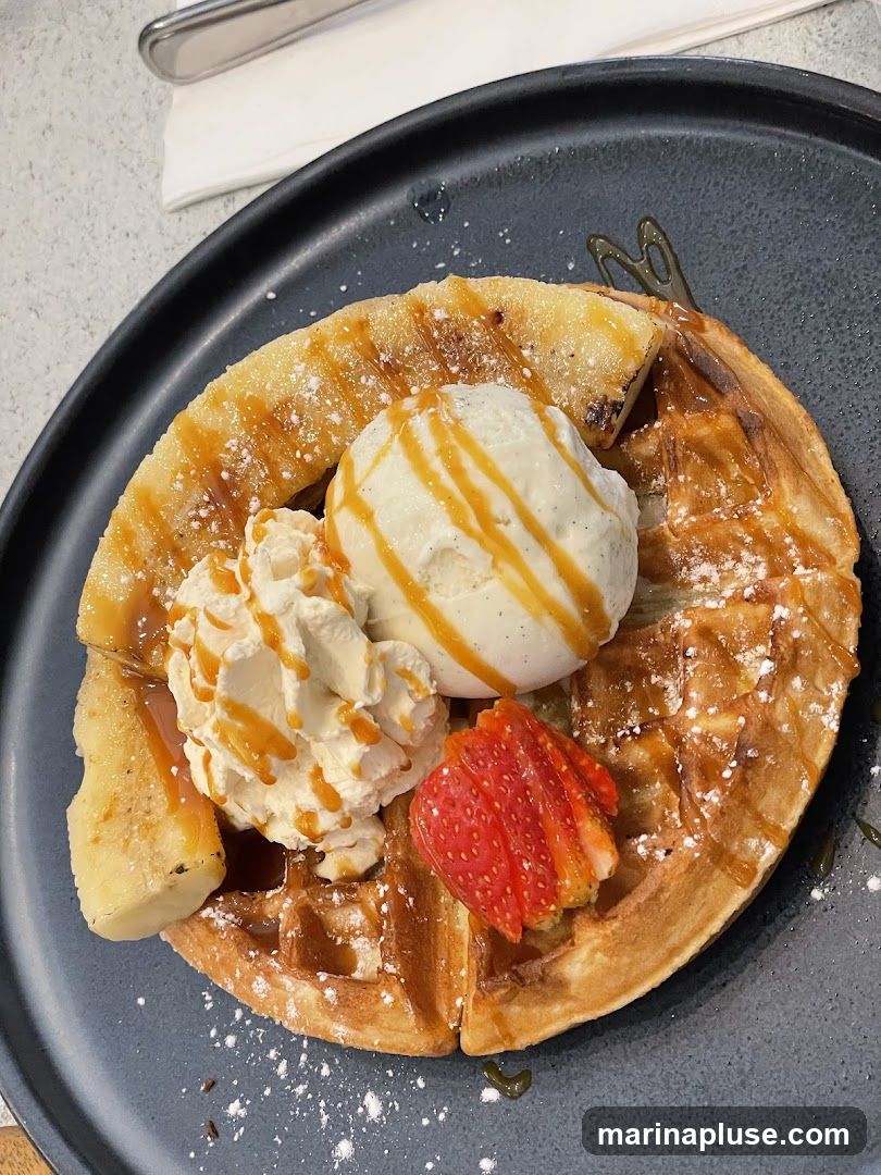 A close-up overhead shot of a golden-brown waffle served on a dark plate. The waffle is topped with a scoop of vanilla ice cream drizzled with caramel sauce, a swirl of whipped cream, sliced grilled bananas, and a strawberry slice. Powdered sugar is dusted over the waffle and toppings.