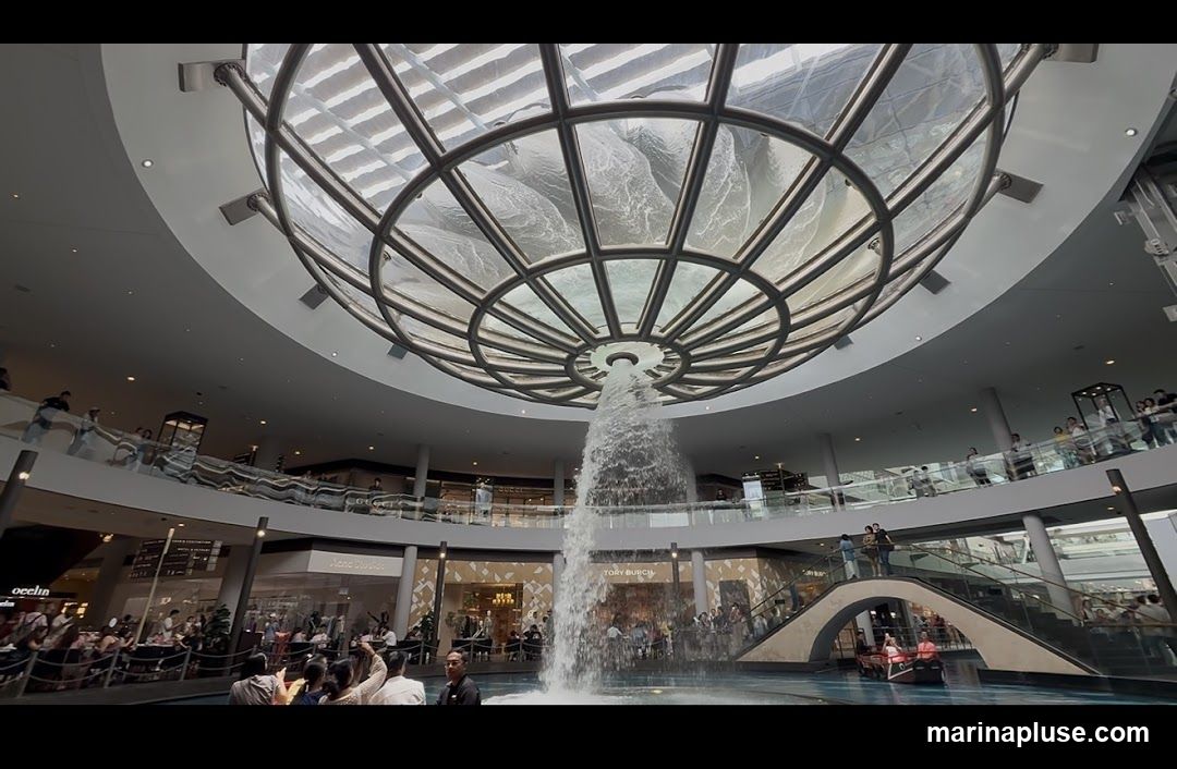 An interior view of a multi-level shopping mall featuring a large, circular skylight with a central water feature cascading down into a pool. People are seen browsing shops and walking on elevated walkways and staircases. Visible store signs include "TORY BURCH" and "Acne Studios". A small boat with passengers is visible in the water.