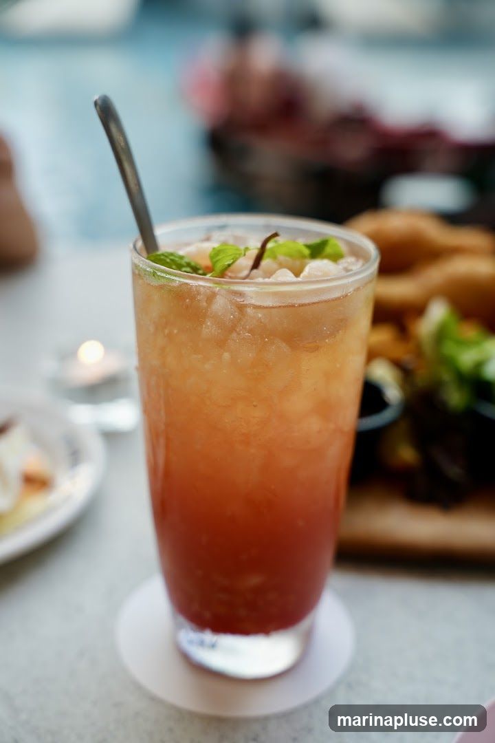 A tall glass filled with a layered orange and red beverage, filled with ice and garnished with mint leaves and what appears to be lychee. A metal spoon is in the glass. In the background, out of focus, are plates of food and a lit candle.