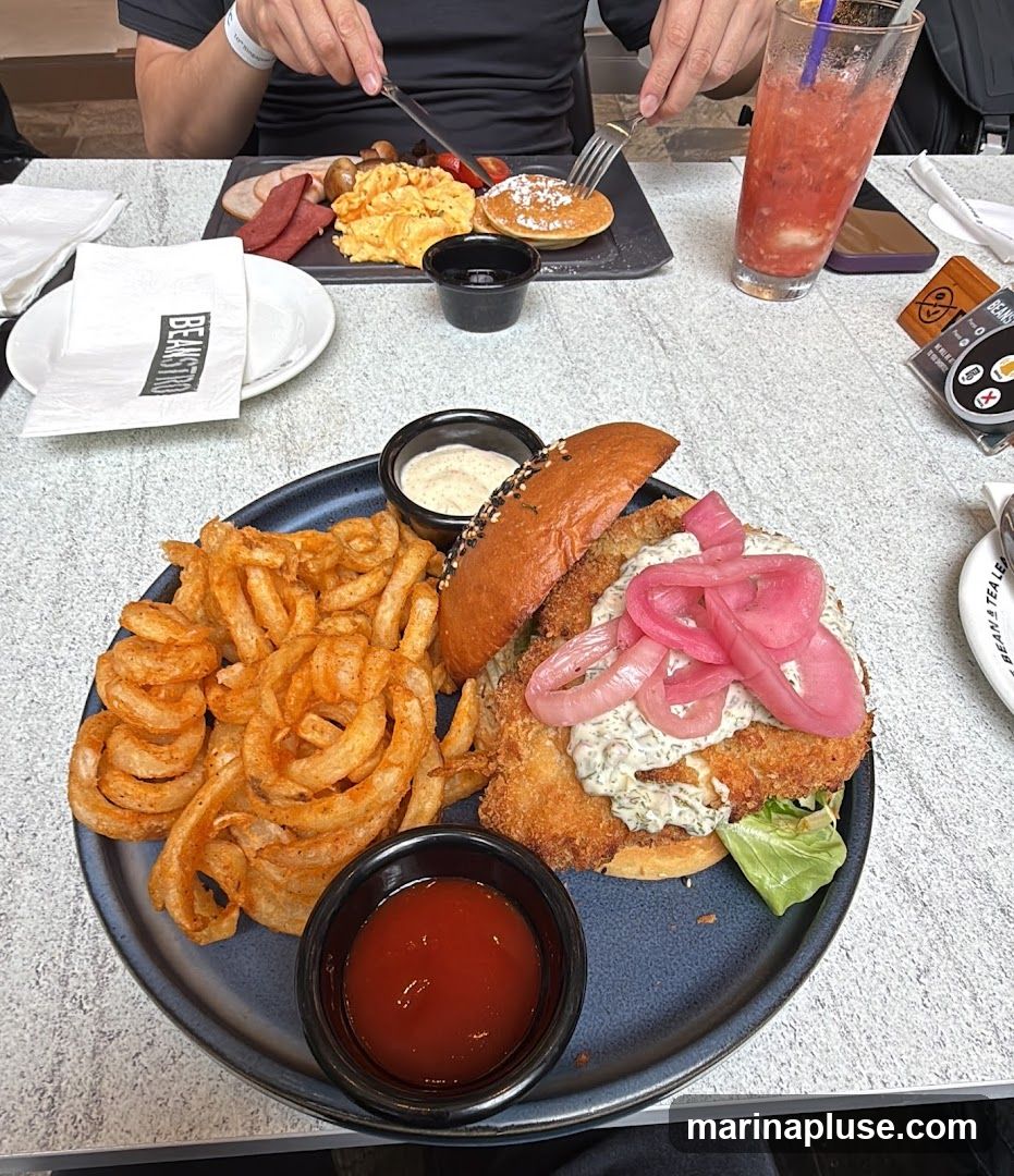A close-up shot of a meal on a table. In the foreground, a large burger with crispy fried chicken, pickled red onions, and a creamy sauce is served with a side of curly fries and ketchup. In the background, a breakfast platter with scrambled eggs, pancakes, and meats is visible, along with a pink beverage in a glass. A napkin with the logo "BEANSTRIO" is also present.