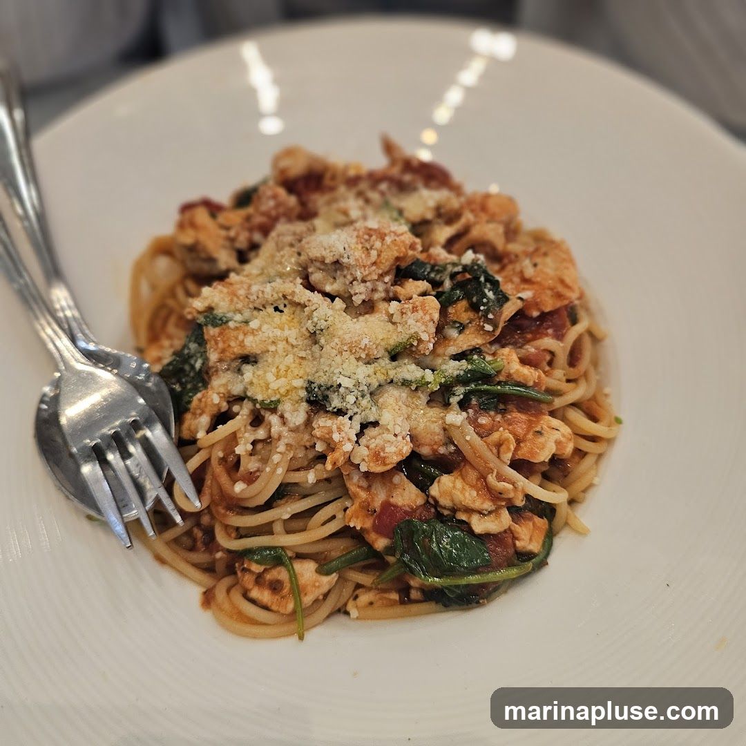 A close-up shot of a plate of spaghetti topped with chicken, spinach, and grated cheese. A fork and spoon are placed on the side of the plate.