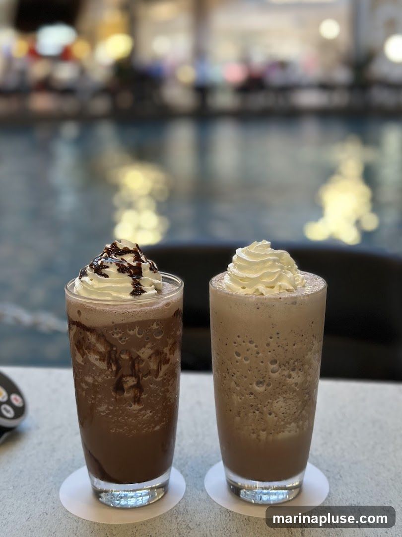 Two tall glasses of chocolate frappes topped with whipped cream sit on a table. The glass on the left has chocolate syrup drizzled on top of the whipped cream and down the sides of the drink. The glass on the right has plain whipped cream. The background is blurred and appears to be an indoor setting with water and lights.