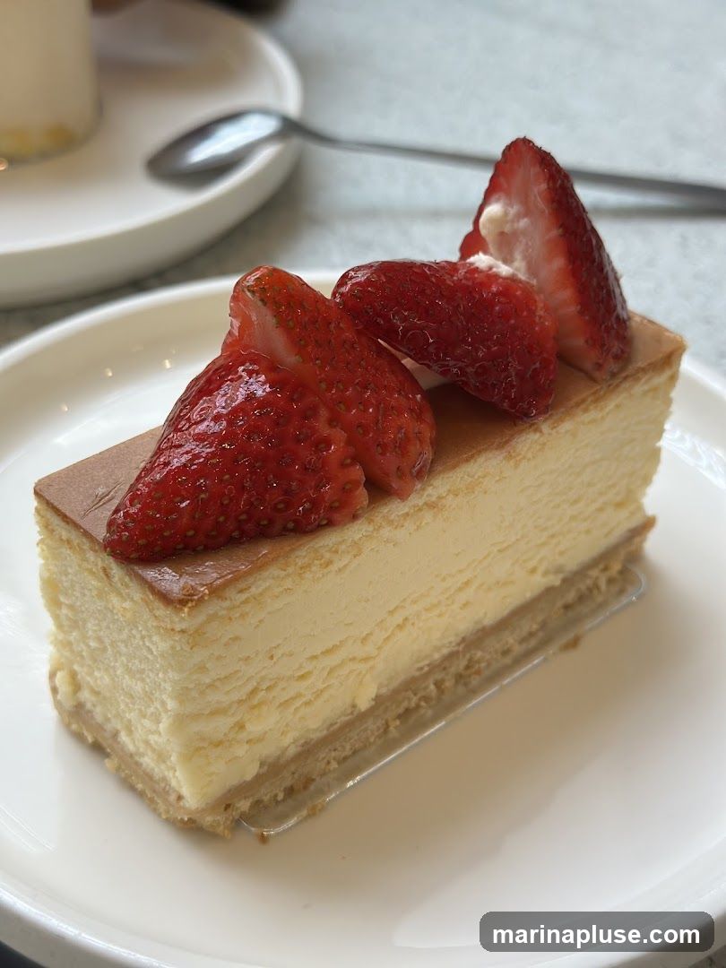 A slice of cheesecake topped with fresh strawberries on a white plate. A spoon and a cup are visible in the blurred background.