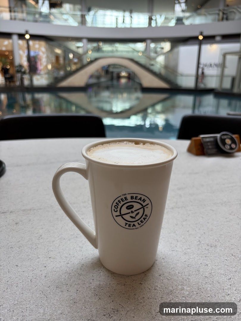 A white mug with the logo of The Coffee Bean & Tea Leaf is filled with a frothy coffee drink. The mug sits on a light-colored, textured surface. In the background, a large indoor space with a water feature and shops is visible.